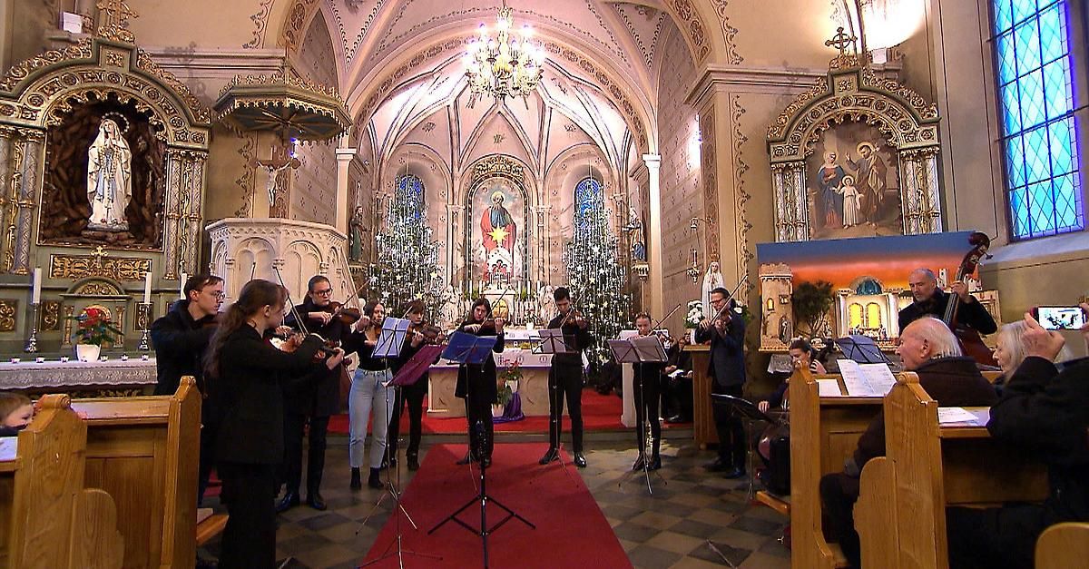 Eine Gruppe von Musikern tritt auf der Bühne in einer Kirche auf. Die Bühne ist mit einem roten Teppich und verschiedenen Musikinstrumenten geschmückt.