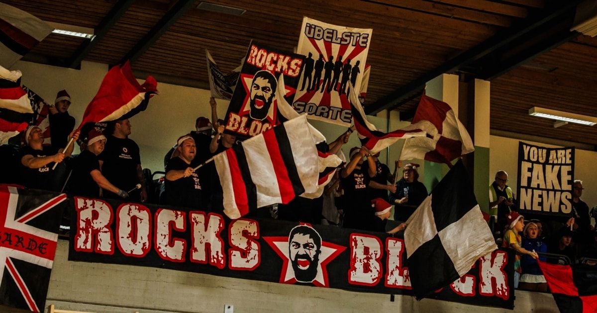 Fans in einem Stadion halten Banner mit den Texten 'ROCKS' und 'BLOOD' und Flaggen. Das Stadion ist mit einem Banner dekoriert, das das Gesicht eines Mannes zeigt.