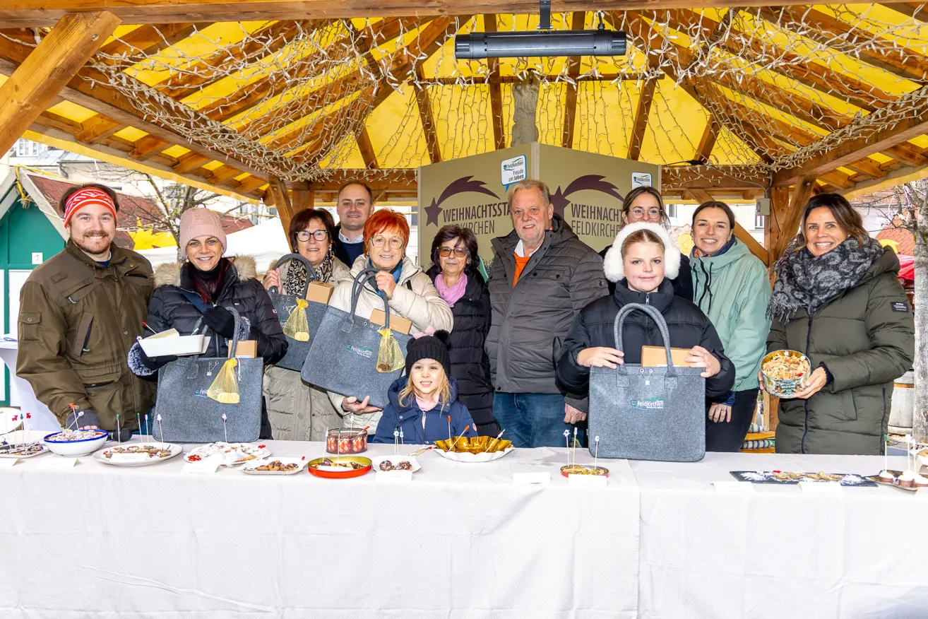 Eine Gruppe von Menschen steht unter einem Pavillon, hält Geschenke und lächelt für ein Foto. Vor ihnen steht ein Tisch mit verschiedenen Speisen. Hinter ihnen befindet sich ein Banner mit den Worten 'Weihnachtskirche'.