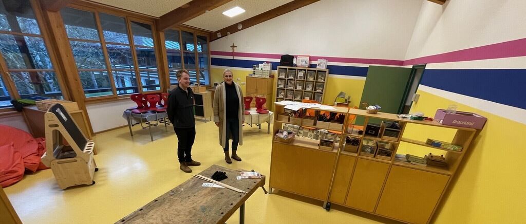 Zwei Personen stehen in einem Raum mit gelbem Boden und Holzschränken, umgeben von Stühlen, Tischen und Regalen. Ein Glasfenster bietet Blick auf das Freie.