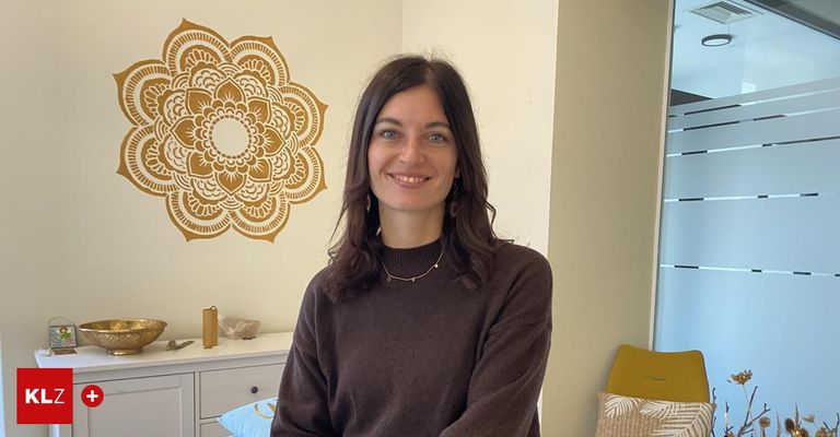 Eine Frau mit braunen Haaren lächelt für ein Foto. Sie trägt einen braunen Pullover und eine Halskette. Sie steht vor einer weißen Wand mit einem dekorativen Mandala. Ein weißer Schrank und ein gelber Stuhl befinden sich neben ihr.