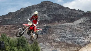 Ein Motocross-Fahrer in roter und weißer Kleidung mit 'LERT' auf dem Rücken ist in der Luft auf einer Dirt-Strecke vor einem Berghintergrund.
