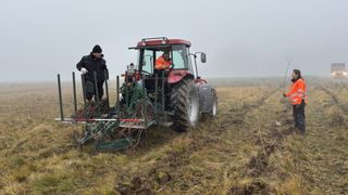 Drei Männer arbeiten auf einem Traktor auf einem Feld. Einer fährt, während zwei andere hinten stehen, einer hält einen Zweig.