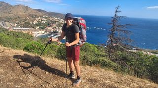 Eine Frau wandert mit einem großen Rucksack auf einem Berg und lächelt für ein Foto, mit einer Küstenstadt und dem Meer im Hintergrund.