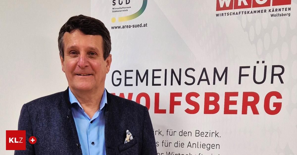 Ein Mann steht lächelnd vor einem weißen Banner mit rotem Text, der 'GEMEINSAM' und 'WOLFSBE' lautet. Er trägt ein blaues Hemd und eine dunkle Jacke.