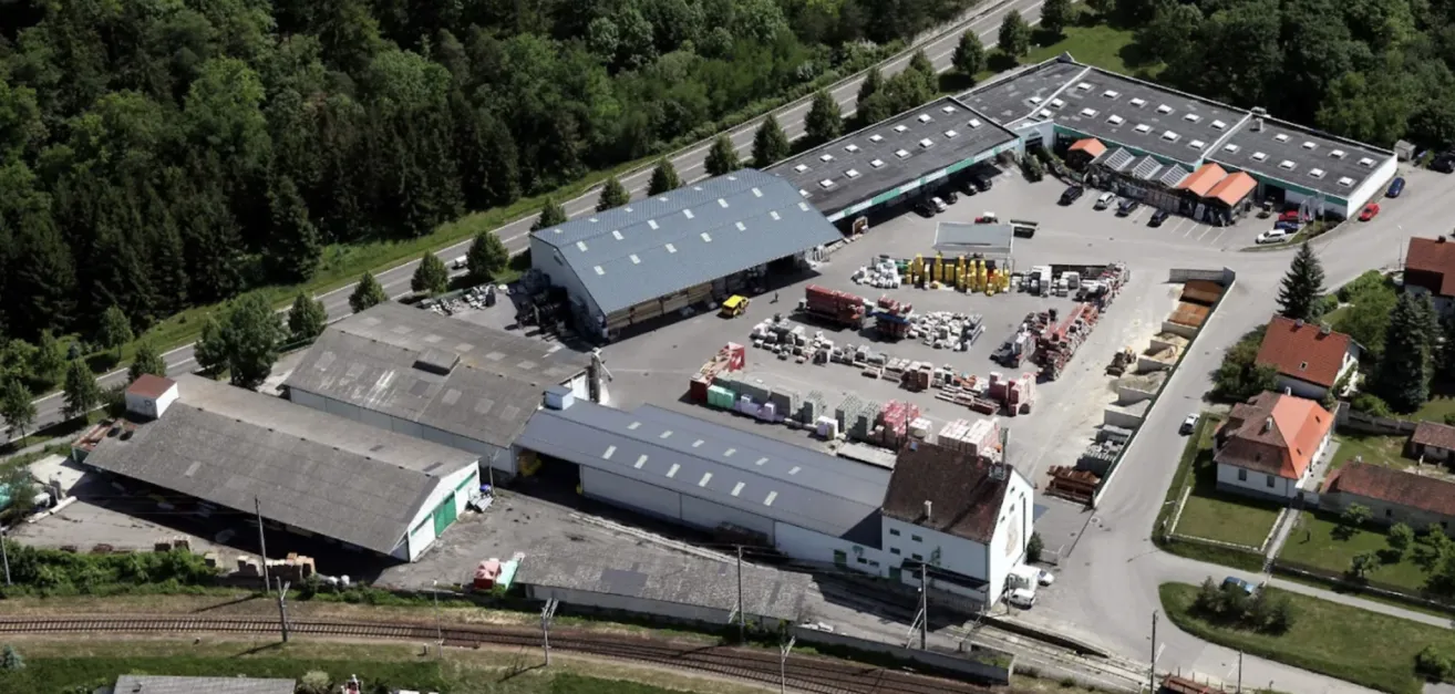 Ein Luftbild eines Industriegebiets mit mehreren Lagerhäusern, einer Straße und geparkten Fahrzeugen. Bäume umgeben das Gebiet, und eine Eisenbahn ist sichtbar.