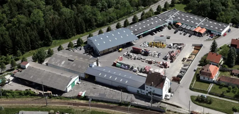 Ein Luftbild eines Industriegebiets mit mehreren Lagerhäusern, einer Straße und geparkten Fahrzeugen. Bäume umgeben das Gebiet, und eine Eisenbahn ist sichtbar.