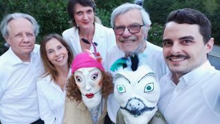 Fünf Personen, darunter drei Frauen und zwei Männer, posieren für ein Foto mit übertriebenen Gesichtsausdrücken. Sie tragen weiße Masken mit bemalten Gesichtszügen.