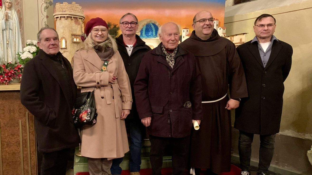 Eine Gruppe von Menschen, darunter zwei Mönche, eine Frau in einem Mantel und zwei Männer, stehen gemeinsam vor einer Kulisse.
