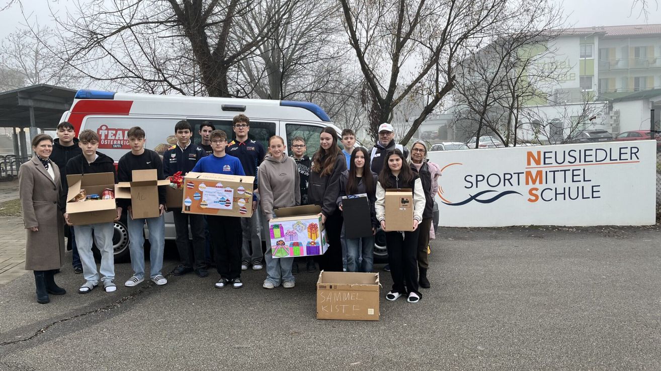 Gruppe von Schülern und Erwachsenen mit dekorierten Kisten vor einem Van und einem Gebäude mit SPORT-Logo.