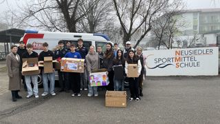 Gruppe von Schülern und Erwachsenen mit dekorierten Kisten vor einem Van und einem Gebäude mit SPORT-Logo.