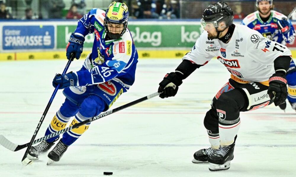 Zwei Eishockeyspieler konkurrieren auf einer Eisbahn. Einer trägt ein blaues Trikot, der andere ein weißes. Beide sind mitten in der Aktion und jagen einem Puck hinterher.