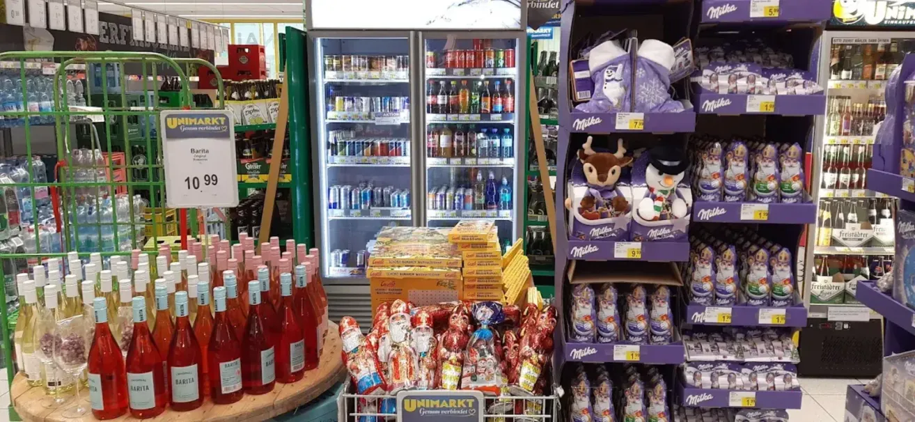 Ein Geschäft zeigt einen Kühler voller Getränke und Snacks mit Weihnachtsmann-Süßigkeiten und festlichen Plüschtieren auf einem Regal. Ein runder Tisch hat Flaschen Wein.