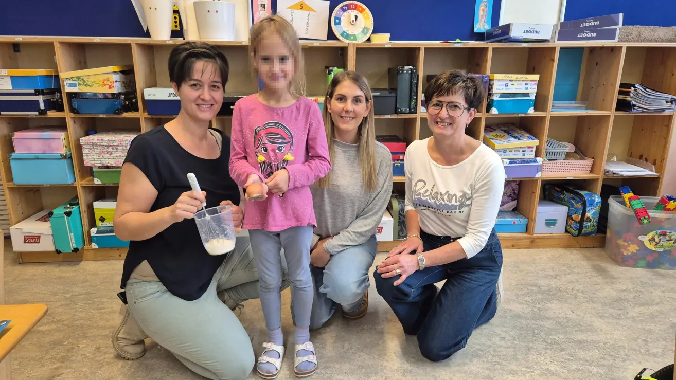 Drei Frauen und ein junges Mädchen posieren lächelnd für ein Foto. Eine Frau hält eine Rührschüssel mit Zutaten. Sie befinden sich in einem Raum mit Regalen und Aufbewahrungsboxen.