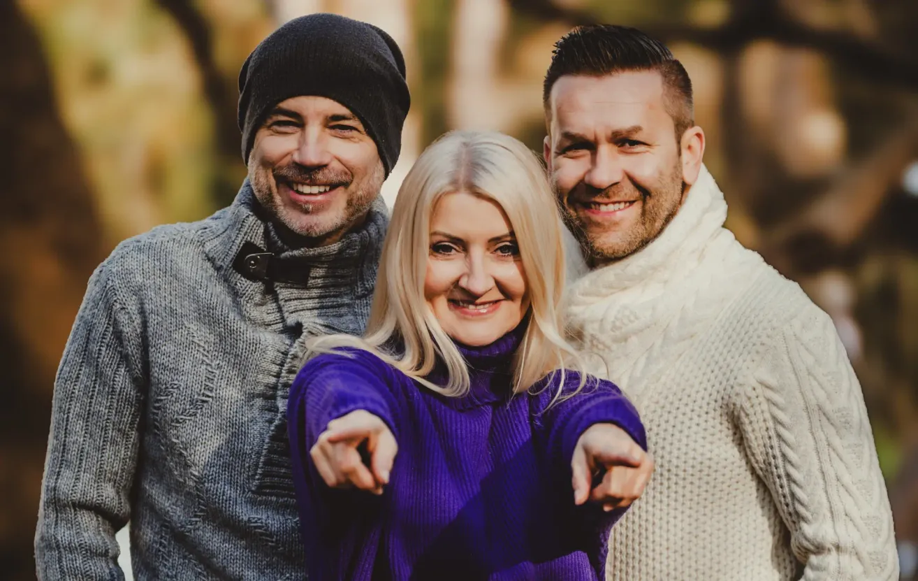 Drei Personen, zwei Männer und eine Frau, lächeln und posieren für ein Foto. Die Frau zeigt mit dem Finger, während die Männer hinter ihr stehen. Sie sind in Winterkleidung gekleidet.