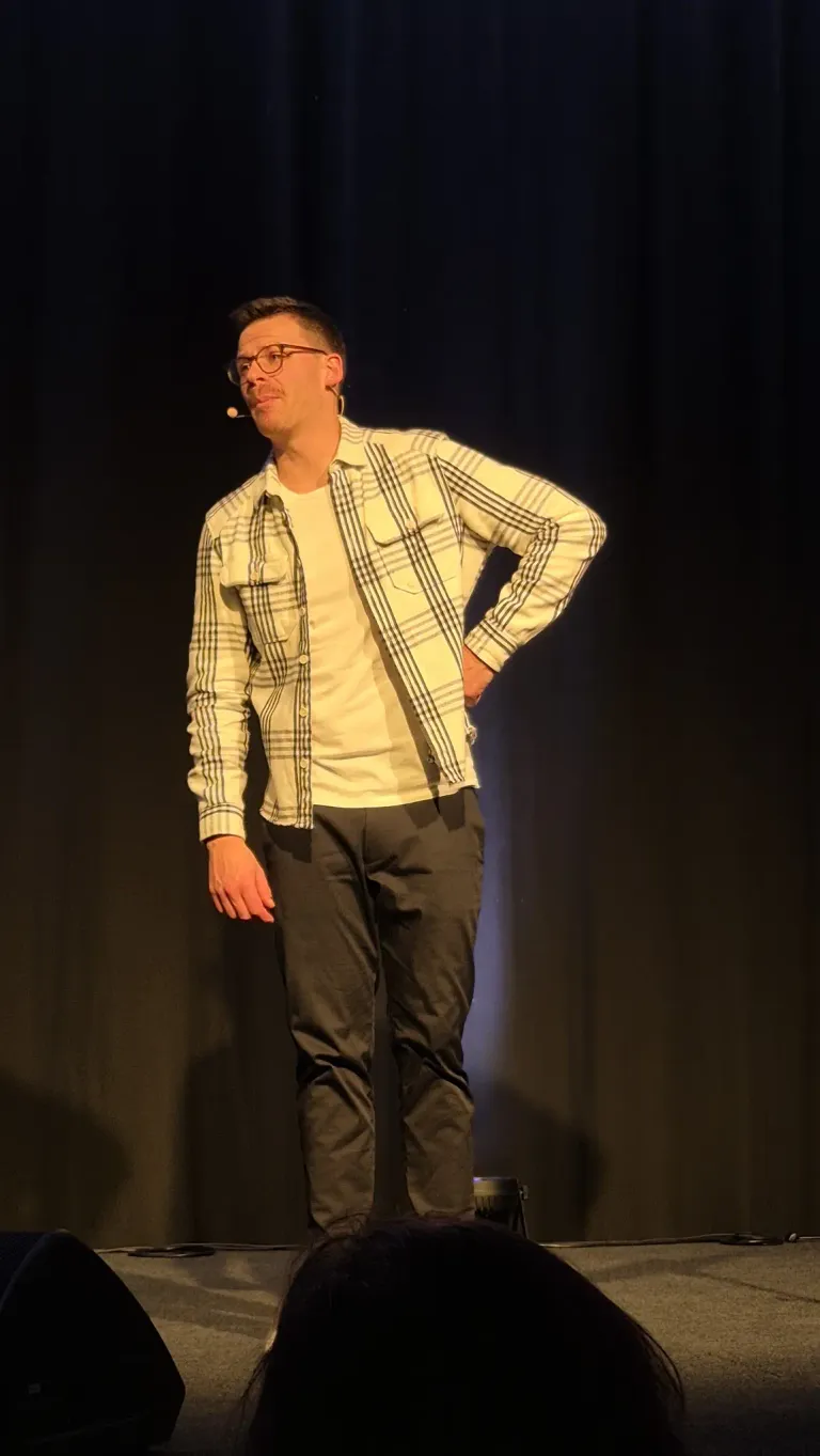 Ein Mann steht auf der Bühne und trägt eine Brille, ein kariertes Hemd und dunkle Hosen. Er hat ein Mikrofon im Ohr und schaut mit den Händen auf den Hüften nach unten.