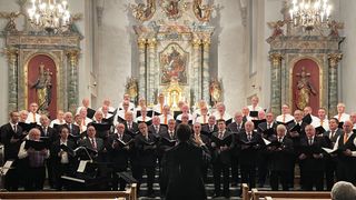 Ein Chor in Anzügen und Krawatten singt in einer Kirche mit aufwendigen Verzierungen, während ein Dirigent vor ihnen steht und die Aufführung leitet.