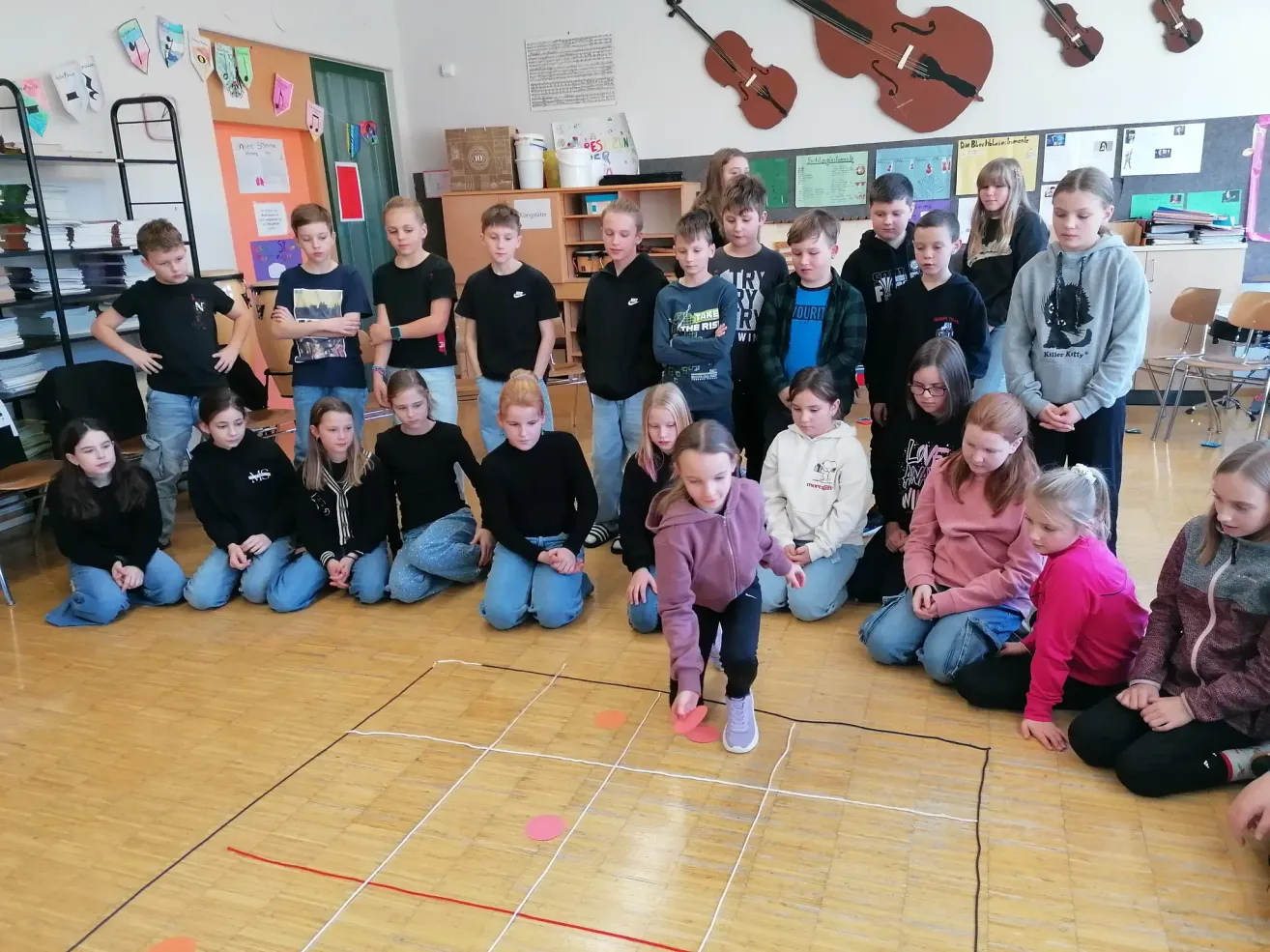 Eine Gruppe von Kindern nimmt an einem Spiel in einem Klassenzimmer teil. Einige sitzen, während andere stehen und zusehen. Musikinstrumente sind an der Wand ausgestellt.