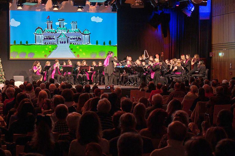 Ein Flötenorchester spielt auf der Bühne mit einer großen Leinwand, die ein Schloss zeigt. Die Musiker sind in Pink und Schwarz gekleidet. Das Publikum sitzt im Vordergrund.