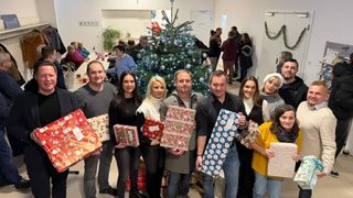 Eine Gruppe von Menschen steht lächelnd und hält Geschenke vor einem geschmückten Weihnachtsbaum. Dahinter sind weitere Personen zu sehen, die plaudern. Die Umgebung ist drinnen, mit einer weißen Wand und einer Tür im Hintergrund.