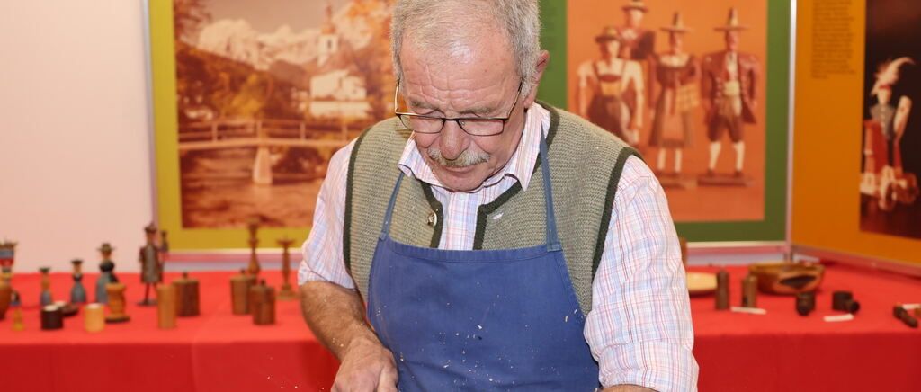 Ein älterer Mann mit Brille und blauer Schürze arbeitet vor einem Tisch mit roter Tischdecke an etwas. Dahinter befindet sich ein verschwommener Hintergrund mit einer Wand und Figuren.
