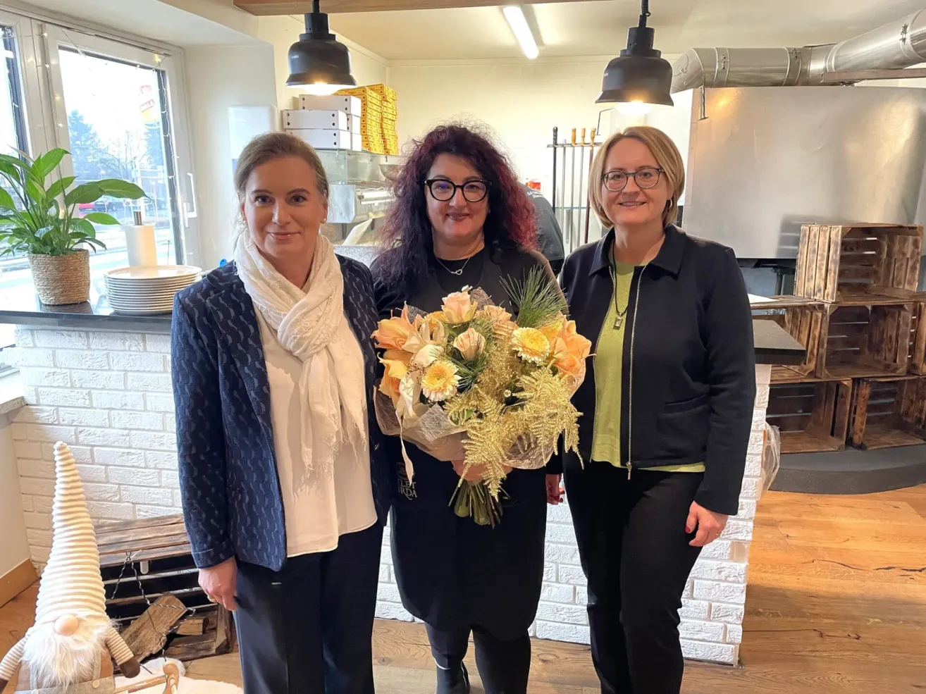 Drei Frauen stehen in einem Raum, eine hält einen Blumenstrauß. Sie lächeln alle und sind in Schwarz gekleidet. Hinter ihnen steht eine weiße Ziegelwand.