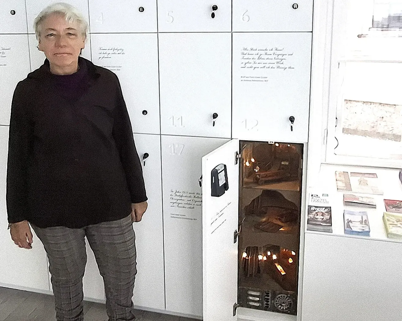 Eine Frau mit weißen Haaren steht neben einem Schließfach mit einem kleinen beleuchteten Fach, das geöffnet ist und eine Kerze und ein Buch zeigt.
