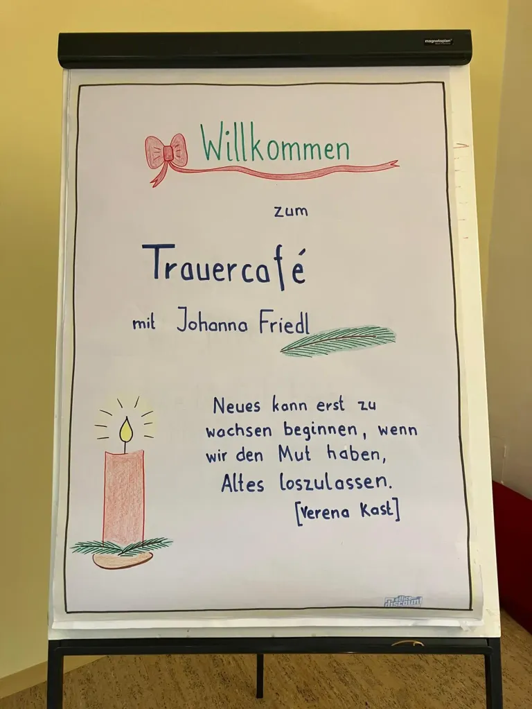 Ein weißes Board mit einer Zeichnung einer Kerze, einer Schleife und einem Blatt, mit der Aufschrift 'Willkommen zum Trauercafe mit Johanna Friedl' und einem Zitat von Verena Kast.