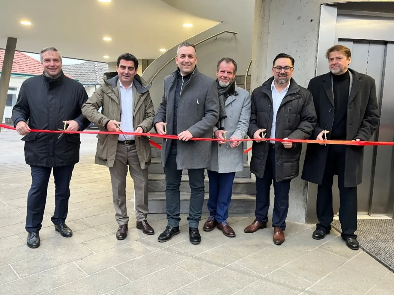 Fünf Männer in Wintermänteln stehen lächelnd vor einem Gebäude, halten ein rotes Band und feiern wahrscheinlich eine Eröffnungsveranstaltung.