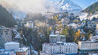 Luftaufnahme einer Stadt mit schneebedeckten Gebäuden, vor dem Hintergrund schneebedeckter Berge und Bäume. Die Stadt liegt in einem Tal, mit einem klaren blauen Himmel darüber.