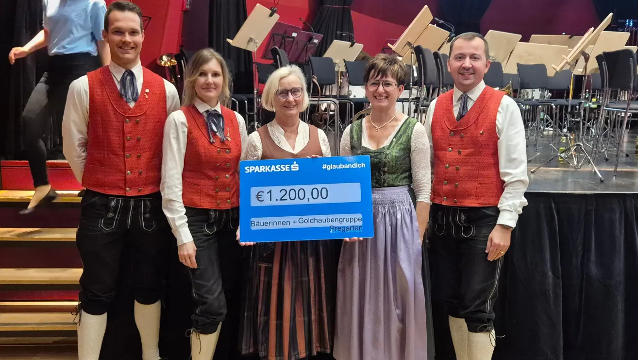 Fünf Menschen in traditioneller Kleidung stehen zusammen, zwei Frauen halten einen Scheck mit einer großen Geldsumme. Die Frauen tragen Brillen und Halsketten. Im Hintergrund sind Musikinstrumente zu sehen.