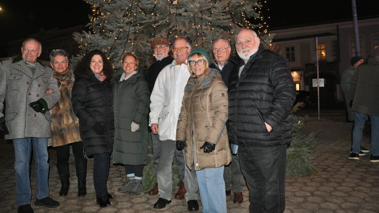 Eine Gruppe von Menschen steht zusammen vor einem geschmückten Weihnachtsbaum bei Nacht. Sie lächeln und scheinen für ein Foto zu posieren.
