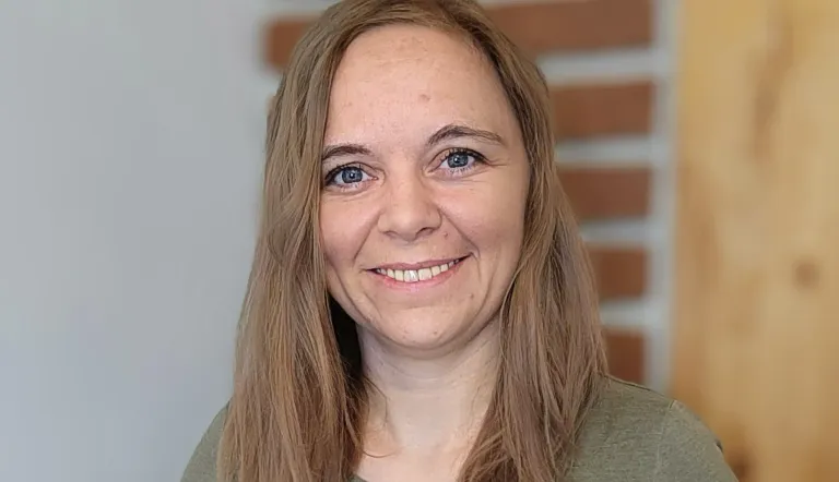 Eine lächelnde Frau mit hellbraunen Haaren und blauen Augen, die ein grünes Hemd trägt, steht vor einer Ziegelsteinwand.