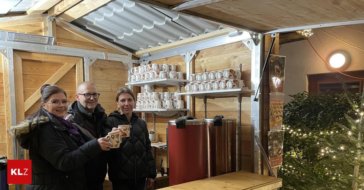 Zwei Personen stehen in einer gemütlichen Hütte und halten Tassen. Hinter ihnen befinden sich Regale voller Tassen und ein Weihnachtsbaum. Sie lächeln und scheinen für ein Foto zu posieren.