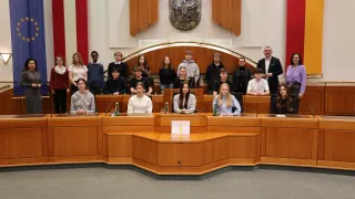 Eine Gruppe junger Menschen in Hoodies und Pullovern sitzt in einem Sitzungssaal, möglicherweise bei einer Ratssitzung. Ein Mann in Anzug steht vor ihnen.