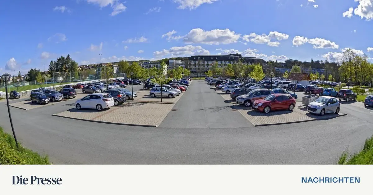 Ein Parkplatz mit vielen Autos in Reihen geparkt, umgeben von Bäumen und Grünflächen. Der Himmel ist blau mit verstreuten Wolken.