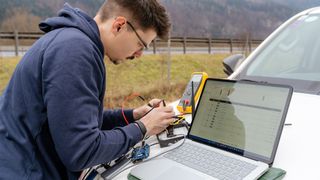 Ein Mann in einer blauen Kapuzenjacke arbeitet an einem Laptop auf einem Auto, mit verschiedenen elektronischen Geräten um ihn herum.