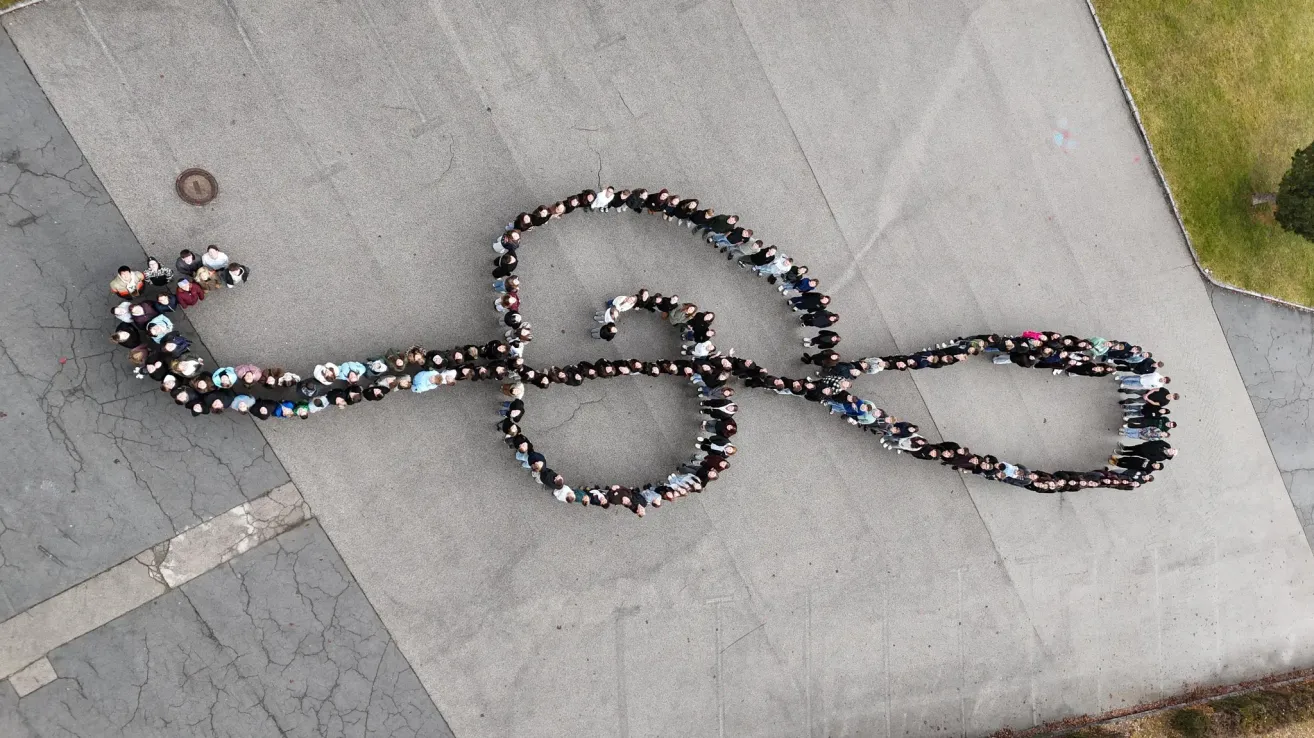 Eine Menschenmenge bildet einen großen Buchstaben 'S' auf einer Betonoberfläche, wobei viele Personen blaue und weiße Kleidung tragen.