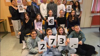 Eine Gruppe junger Menschen in einem Klassenzimmer, die Schilder mit Zahlen und dem Euro-Symbol halten. Einige haben Herzen an der Wand hinter ihnen gemalt.
