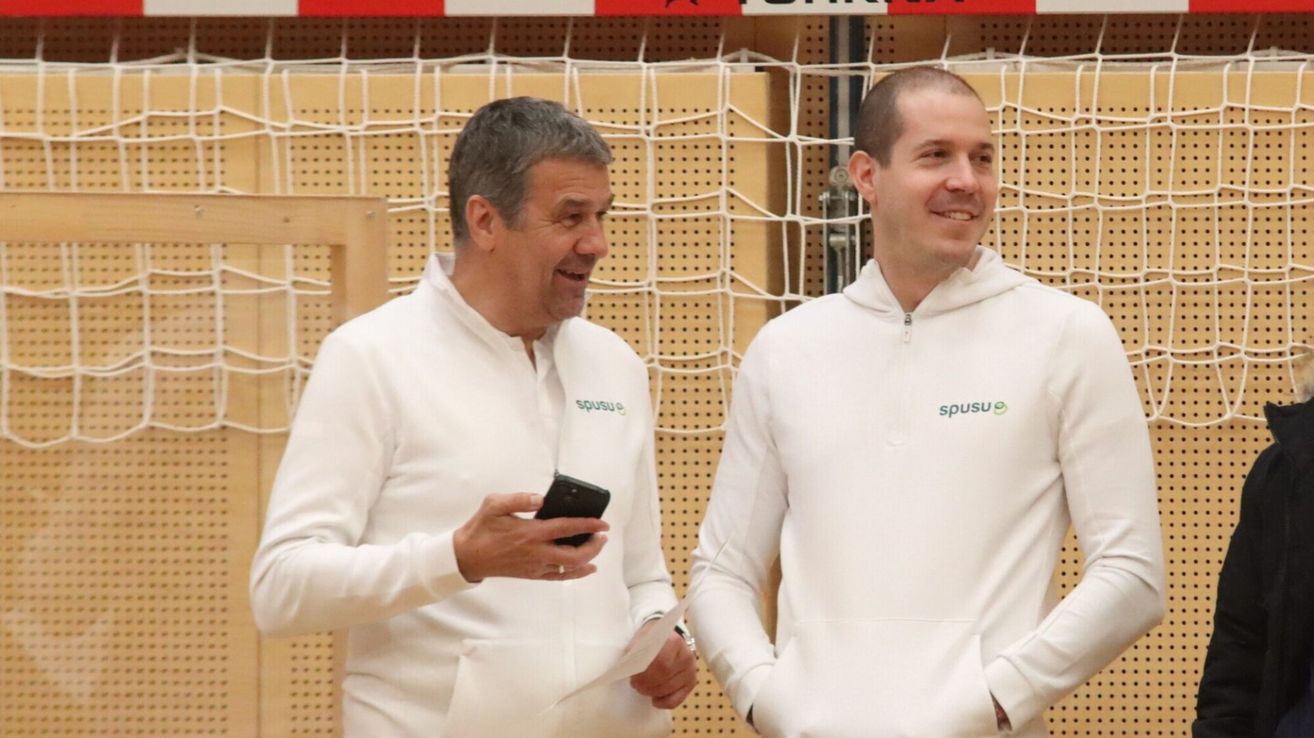 Zwei Männer in weißen Hoodies mit dem Logo 'spu' stehen nebeneinander und lächeln. Einer von ihnen hält ein Handy und scheint etwas darauf zu zeigen.