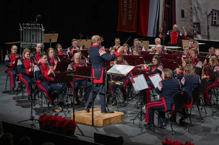 Ein Militärorchester spielt auf der Bühne mit roten Blumen vorne. Der Dirigent steht in der Mitte und die Musiker sitzen. Ein roter Banner mit weißen Text steht hinter dem Orchester.
