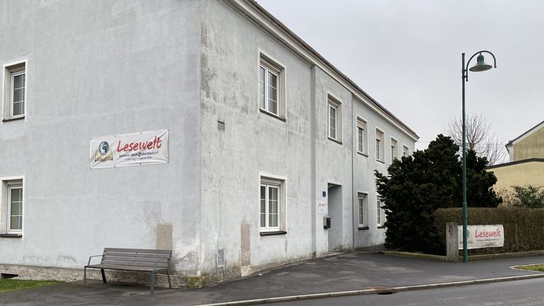 Ein graues Gebäude mit einem Schild, das 'Lesewelt' auf der Wand anzeigt. Das Gebäude hat mehrere Fenster und eine Tür. Es gibt eine Bank und einen Baum auf der rechten Seite. Die Straße ist asphaltiert.