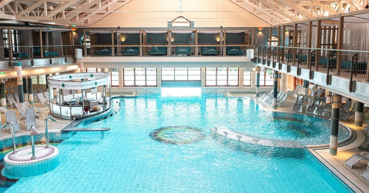 Ein großer Innenpool mit blauem Wasser und gefliesten Böden. Es gibt Liegestühle an der Seite und eine zweite Etage mit mehreren Stühlen und Tischen.