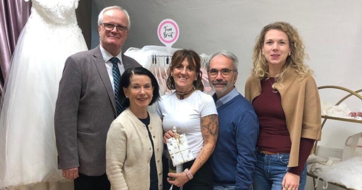 Fünf Menschen stehen nebeneinander und lächeln. Eine Frau in der Mitte hält ein verpacktes Geschenk. Hinter ihnen ist ein Brautkleid an einem Kleiderbügel.