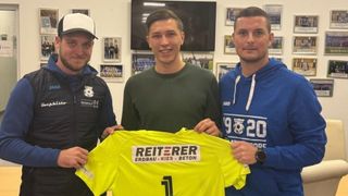 Drei Männer posieren mit einem gelben Trikot. Der Mann in der Mitte lächelt und hält das Trikot. Der Mann auf der linken Seite trägt eine Kappe. Der Mann auf der rechten Seite trägt eine Uhr.