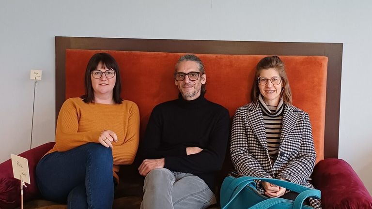 Drei Personen sitzen zusammen auf einer Bank und lächeln alle. Die Frau auf der linken Seite trägt eine Brille und ein orangefarbenes Oberteil. Der Mann in der Mitte trägt eine Brille und ein schwarzes Rollkragenhemd. Die Frau auf der rechten Seite trägt einen karierten Mantel und ein gestreiftes Oberteil.