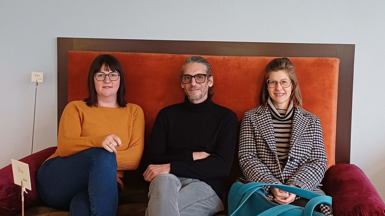 Drei Personen sitzen zusammen auf einer Bank und lächeln alle. Die Frau auf der linken Seite trägt eine Brille und ein orangefarbenes Oberteil. Der Mann in der Mitte trägt eine Brille und ein schwarzes Rollkragenhemd. Die Frau auf der rechten Seite trägt einen karierten Mantel und ein gestreiftes Oberteil.