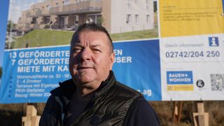 Ein Mann steht vor einem großen blauen Schild, mit einem Gebäude und einem Balkon im Hintergrund.