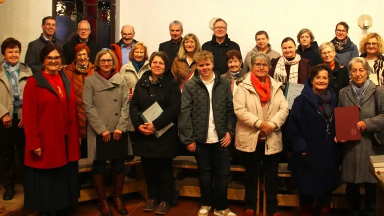 Eine Gruppe von Menschen steht vor einer weißen Wand, einige tragen Brillen, Schals und Stiefel. Sie halten Urkunden in der Hand und posieren für ein Foto.