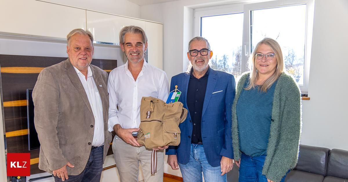 Vier Menschen stehen eng beieinander in einem hellen Raum mit einem hellen Fenster und lächeln für ein Foto. Ein Mann in einem weißen Hemd hält eine braune Tasche. Hinter ihnen zeigt eine Wand und ein Glasfenster einen Blick auf Bäume.